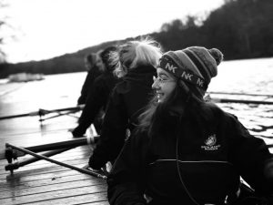 Gabriela sits in a racing shell by a dock. There are some people behind them and they are looking toward the left side of the photo, wearing a hat. The photo is in black and white.