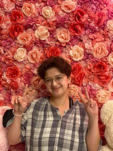 Gabriela pictured with a smile, grey and white blouse, making a peace sign with each hand. The background is composed of flowers in various shades of pink.